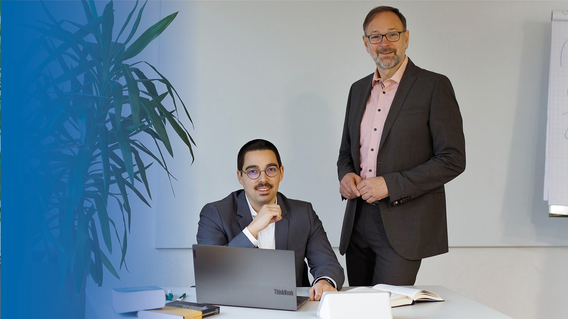 Die Rechtsanwälte Herr Kröll sitzend und Herr Weber stehend in Anzügen vor einem Büroschreibtisch zur Kamera lächelnd.